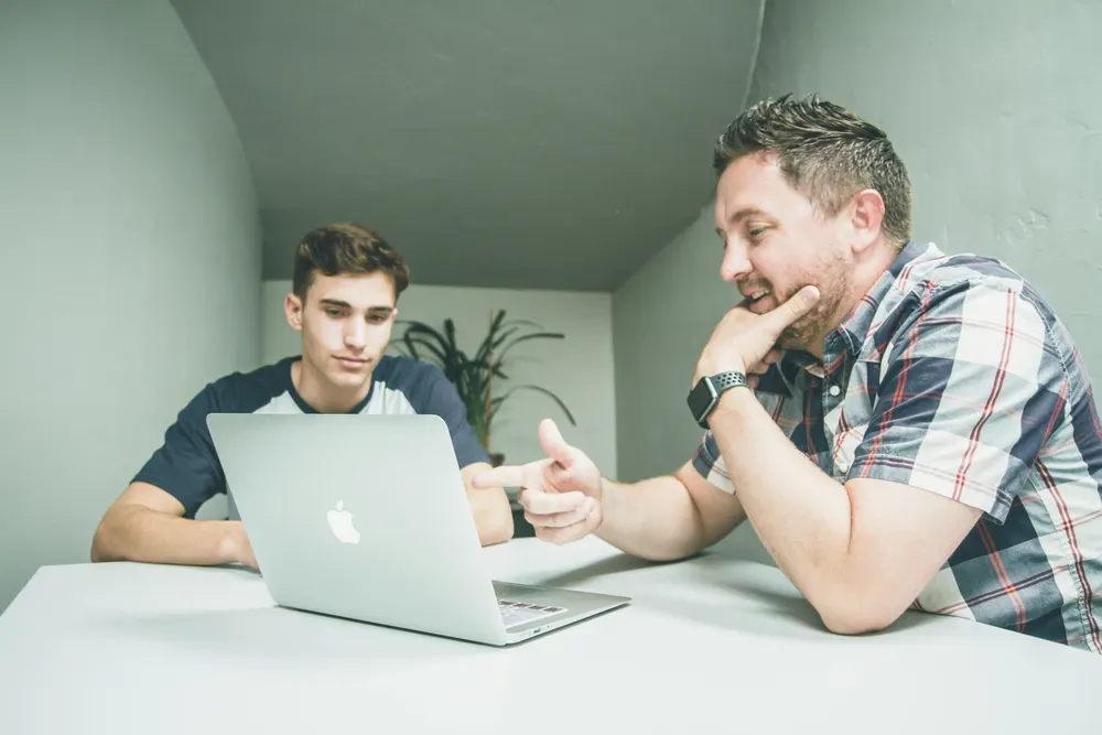 One man points at a laptop screen while another looks on