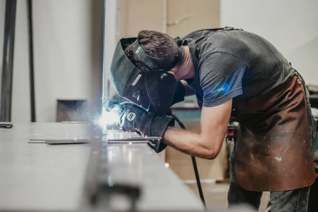 A welder in a helmet working at a bench in a metal shop