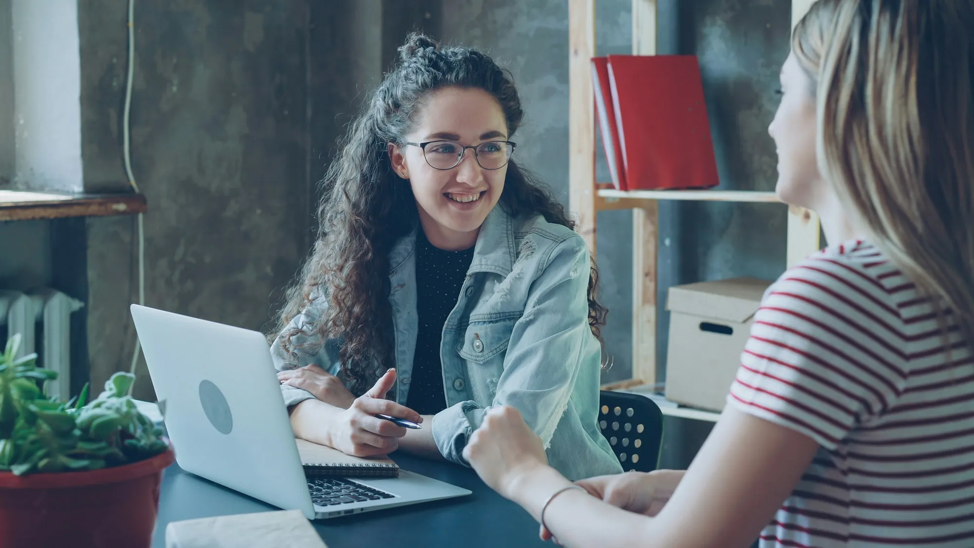 Two women talk and laugh across a desk with a laptop